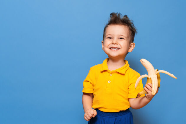 快乐一个穿着黄色t恤的孩子拿着剥了皮的香蕉当食物的快乐肖像 摄影棚拍摄隔离快乐香蕉快乐