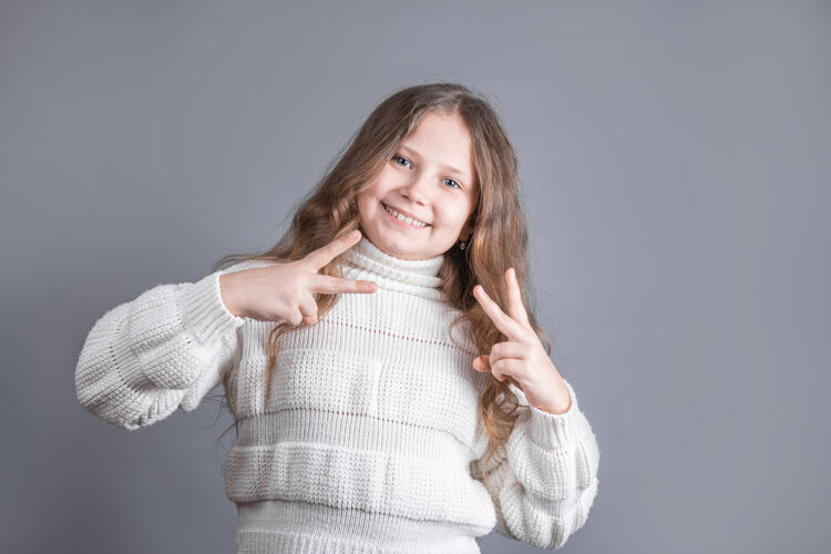 孩子一个年轻迷人的金发小女孩的肖像 穿着毛衣 在灰色的工作室背景上 双手都有v字标志 和平标志 胜利手势学校时尚美丽
