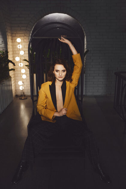 裤子性感的年轻女子穿着黄色外套 赤裸着身体 穿着时髦的格子裤 坐在一把金属椅子上 在黑暗的室内摆姿势形状色情成人