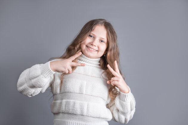 孩子一个年轻迷人的金发小女孩的肖像 穿着毛衣 在灰色的工作室背景上 双手都有v字标志 和平标志 胜利手势学校时尚美丽