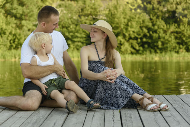 户外幸福的小家庭 爸爸妈妈和小金发儿子坐在码头上一起太阳成人