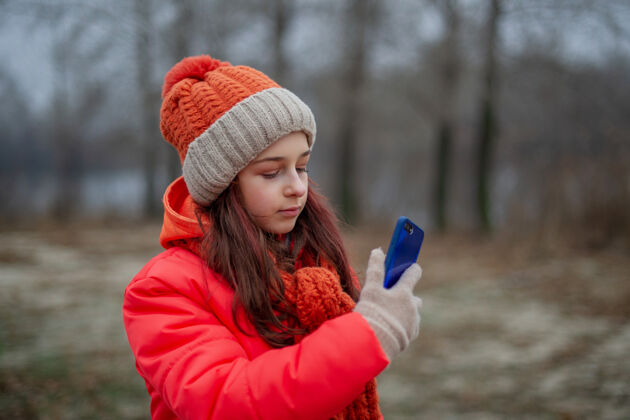 户外一个身穿橙色夹克 戴着帽子 戴着围巾 手里拿着智能手机的少女正在自拍漂亮使用人