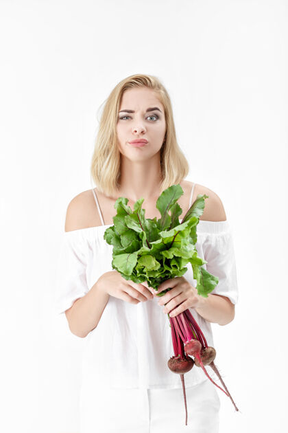 健康心怀不满的金发女人穿着白色上衣 手里拿着一根长着绿叶的甜菜根背景.健康还有维生素肖像蔬菜绿色