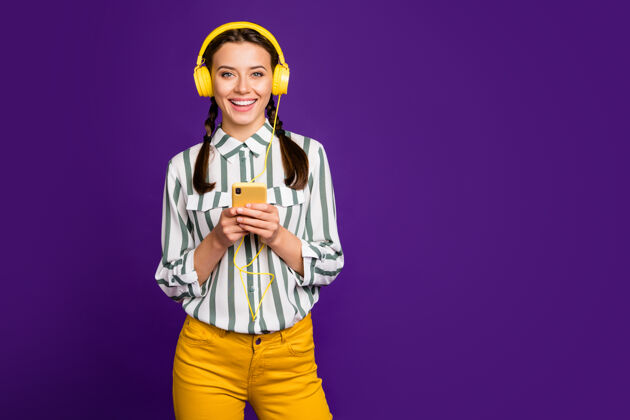辫子照片中美丽的年轻女士手持电话积极度假时间听音乐穿凉爽的耳罩条纹衬衫黄色裤子孤立的紫色背景旋律女性兴奋