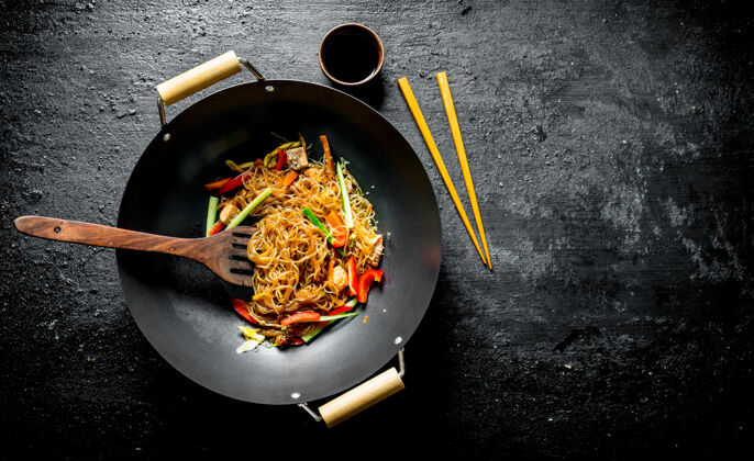 猪肉中国玻璃纸意大利面 平底锅 锅铲和筷子.on黑色乡村餐桌传统素食蔬菜