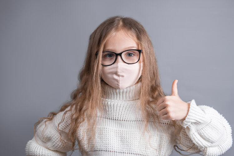 流感一个穿着白色毛衣 戴着医用防护面具的年轻漂亮小女孩的肖像向上竖起大拇指孩子手势症状