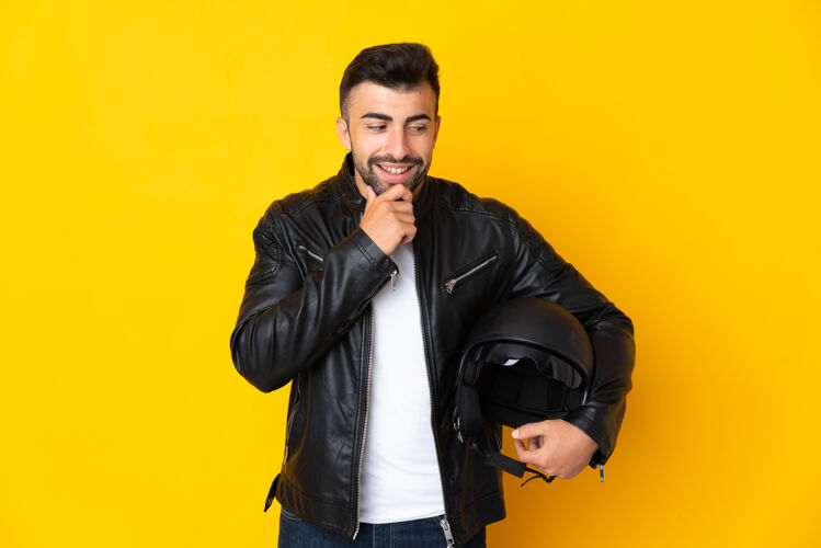 自行车一个戴着摩托车头盔的白人男子站在孤立的黄色背景下 面带微笑地看着旁边拉丁语驾驶肖像