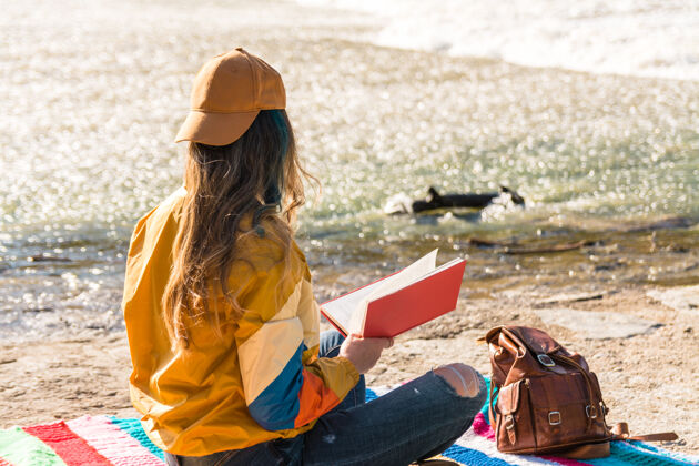 运动戴着帽子 太阳镜 金色运动夹克 皮背包和绿色眼镜的女孩坐在路边五颜六色的毯子上河阅读一本书自然放松时间生活方式概念橱柜女士态度