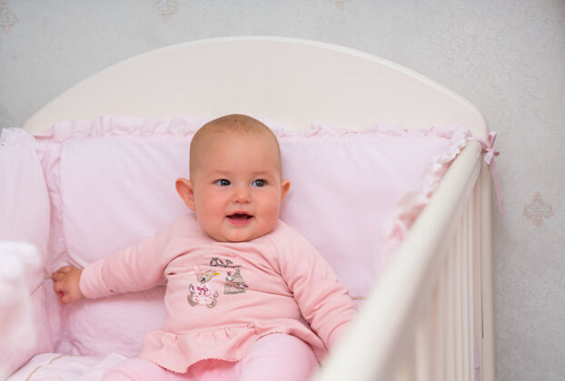 关注小女婴坐在粉红色的婴儿床上 穿着可爱的粉色套装 对着镜头微笑玩具移动的智力