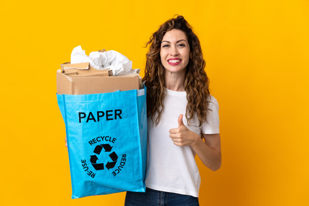 好年轻女子拿着一个装满废纸的回收袋在黄色上做着竖起大拇指的手势保护手指拇指