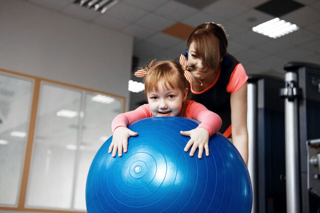 运动穿着粉色上衣的小女孩和妈妈一起玩健身球 妈妈在健身房帮女儿跳健身球 快乐的女孩正在玩健身球体操锻炼健身房