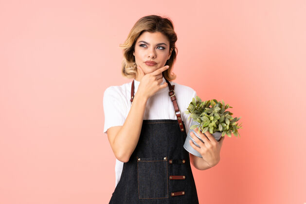 看俄罗斯园丁女孩抱着一株孤立的粉红色植物在想一个主意怀疑女人观念