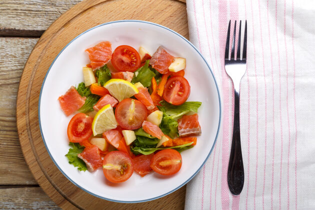 新鲜沙拉 三文鱼和樱桃番茄和绿色沙拉在一个盘子里放在一个木制的桌子上 旁边放着叉子和餐巾鳟鱼三文鱼沙拉菠菜