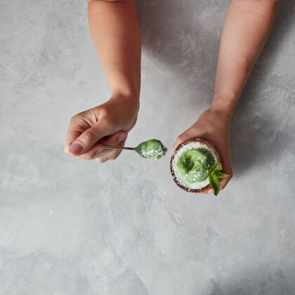 刷新美味的绿色甜点 冰激凌在椰子壳里 女人手里拿着勺子放在灰色的水泥桌上空格.top查看素食生食贝壳
