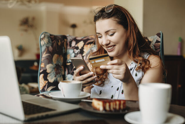 手机迷人的大块头红发女人手持信用卡和智能手机 坐在咖啡馆里用笔记本电脑进行在线交易在线成人饮食