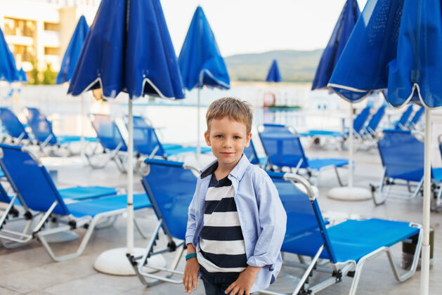海岸线在一个避暑胜地 一个小男孩站在蓝色雨伞和日光浴者中间奢华肖像欢呼