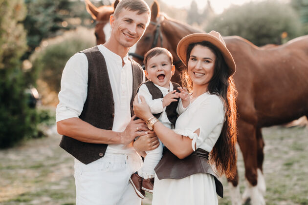 骑马一个穿着白色衣服的家庭和他们的儿子站在大自然中两匹美丽的马旁边一对时髦的夫妇带着一个孩子与马合影白布休闲夜晚