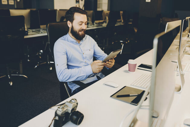 欢呼迷人的胡须男子微笑着浏览现代平板电脑 坐在办公桌旁办公室愉快的办公室里用平板电脑的家伙胡须职业连接