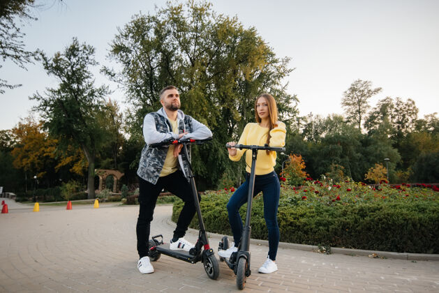 情侣在一个温暖的秋天 一对年轻漂亮的夫妇在公园里骑着电动滑板车乐趣美丽踢滑板车