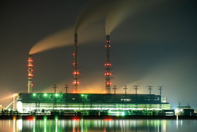 气体灯光明亮的燃煤发电厂高高的管道 黑烟在夜间向上移动 污染了大气 反射在湖水中全球变暖温室塔