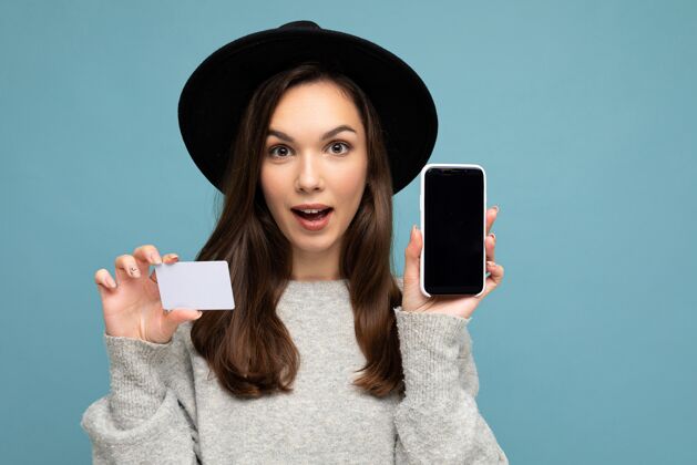 显示一个戴着黑帽子 穿灰色毛衣的女人隔着蓝色的墙 手里拿着信用卡和空着显示屏的手机电话年轻电话