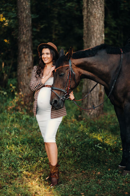 怀孕一个大肚子的怀孕女孩戴着帽子在森林里的马旁边自然时尚穿白色衣服和棕色夹克的女孩大肚子父母马