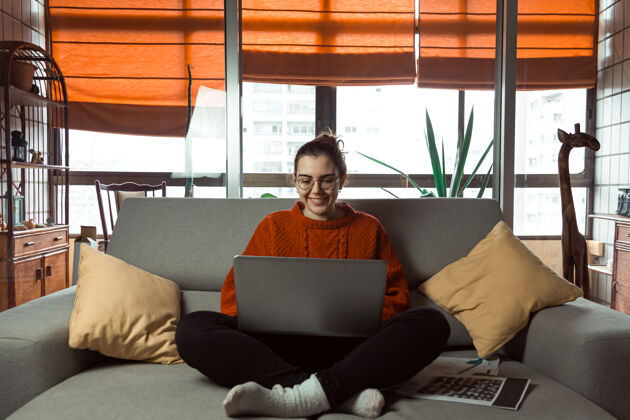 起居一个戴着眼镜的年轻女子一边微笑 一边在沙发上的笔记本电脑上工作和学习文件女性笔记本电脑