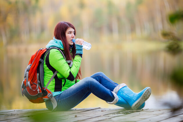 背包年轻的黑发女人在树林里的池塘里用瓶子喝水孤独可爱水