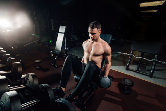 锻炼帅哥赤膊男子举起沉重的哑铃 而坐在长凳上 在黑暗的健身房健康训练肌肉