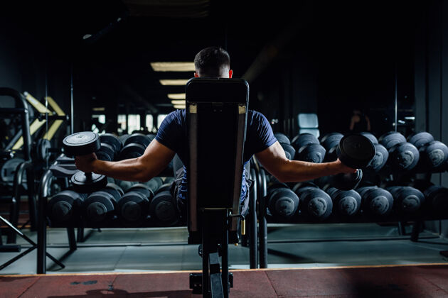 健身房健美运动员在黑暗的健身房里用哑铃练习肱二头肌和坐姿手臂物理家伙