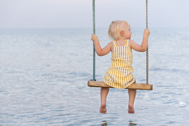 海岸穿着黄色连衣裙的金发女孩坐在一根绳子上水秋千在海上美丽度假休闲