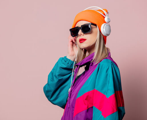 90年代时尚女孩在80年代的运动服和太阳镜与耳机在粉红色的墙上科技Dj设备