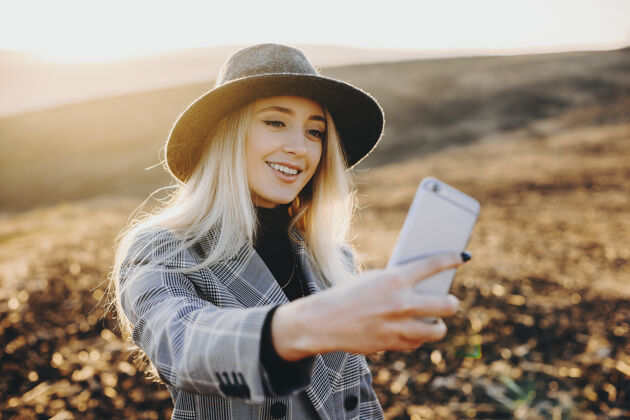 金发美丽的金发白种女人戴着帽子在美丽的日落下用智能手机自拍女性风景日落