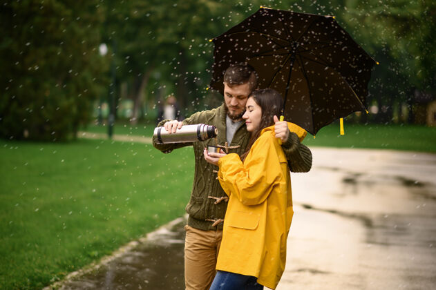 保护情侣在公园喝热茶 夏天下雨戴曼雨中的女人站在雨伞下 漫步小径上的浪漫约会 小巷里的潮湿天气人类丈夫人