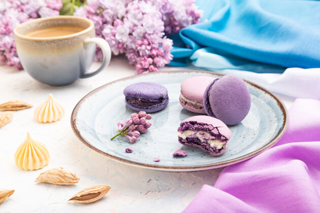 成分紫色的马卡龙或马卡龙蛋糕和一杯咖啡放在白色的混凝土表面和品红蓝色的纺织品上传统蛋白酥皮魅力
