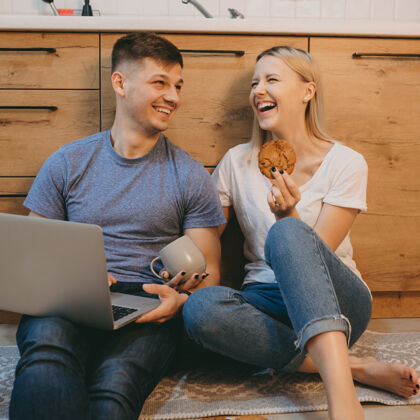 微笑一对迷人的年轻夫妇 早上坐在厨房的地板上笑着 男人在笔记本电脑前工作 女人在吃饼干舒适休闲女朋友
