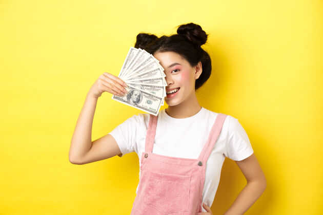 温柔购物漂亮亚洲女模特 化着迷人的妆容 用钞票遮住半张脸 对着镜头性感地微笑 黄色女性金钱美元