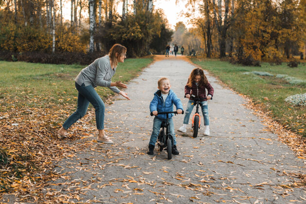 牙齿美丽的年轻家庭在公园里户外玩耍 女儿和儿子骑着自行车 母亲在开玩笑小快乐教学