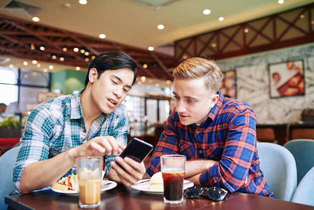 男人亚洲年轻人在咖啡馆和朋友共进午餐 并向朋友展示智能手机上的新手机应用程序分享手机搜索