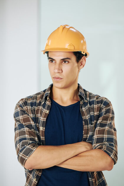 施工英俊严肃的年轻混血青年的画像戴着安全帽的年轻建筑工人交叉双臂 望向别处室内设备业务