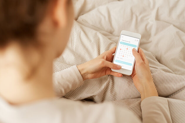 移动年轻女子坐在床上 计划在网上日历她的工作日的后视图早晨舒适购买