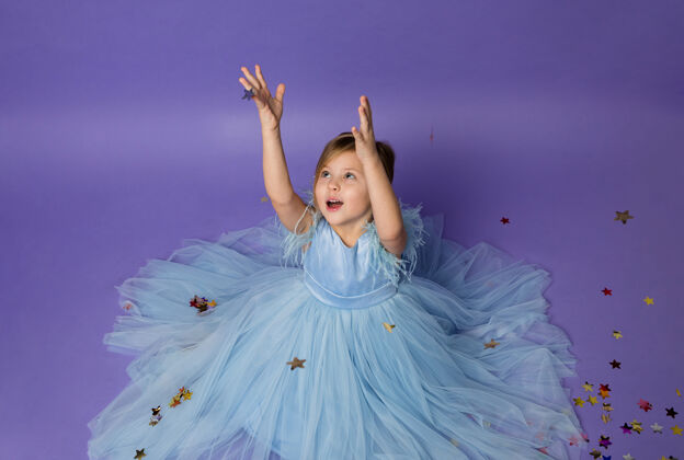 表情穿着蓬松的蓝色连衣裙的小女孩用手抓住紫色的五彩纸屑脸欢乐小