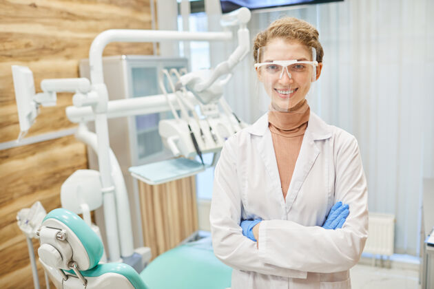 欢呼身穿白大褂 戴着护目镜 双臂交叉 面带微笑地站在诊所前面的年轻牙医的画像微笑肖像快乐