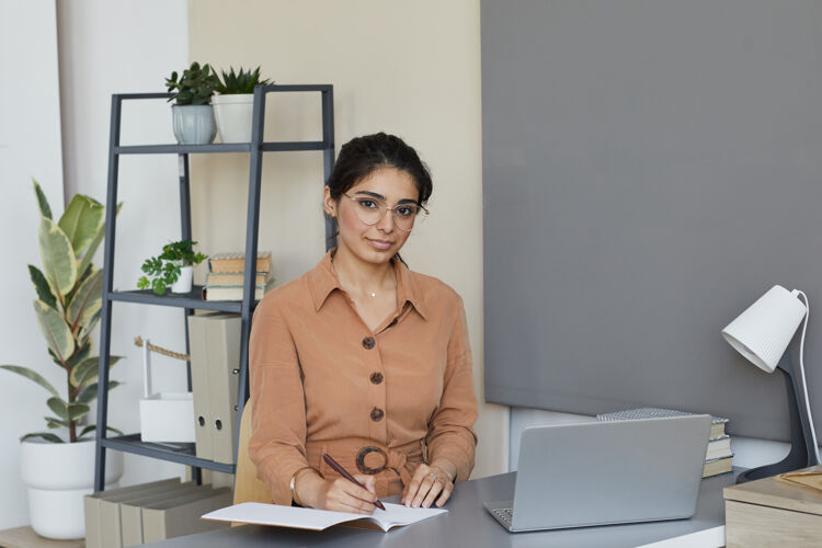 成功一幅年轻女商人坐在办公室里拿着笔记本电脑在笔记本上做笔记的画像她在办公室工作办公室计算机桌子