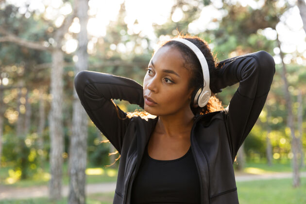 自然20多岁的女跑步者穿着黑色运动服 戴着耳机 穿过绿色公园跑步私人教练运动