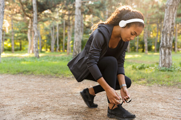 健身20多岁的女运动员穿着黑色运动服系鞋带 在绿色公园里奔跑户外吸引力伸展