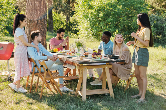 桌子夏日 年轻的朋友们在松树下的自然环境中 坐在餐桌旁聊天后代人吃饭