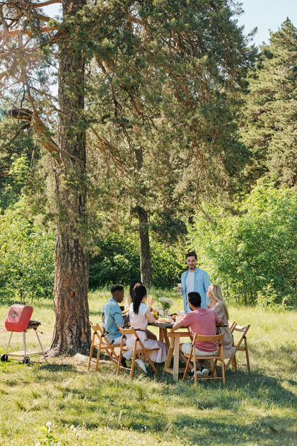 年轻人在夏天的周末 站在旁边的年轻人在松树下的户外晚餐上为他的朋友们端上桌子 并念祝酒词草自然欢呼