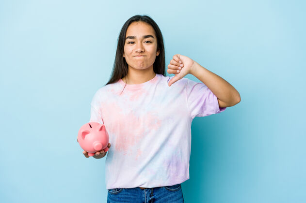 拇指一位年轻的亚洲女士拿着一个粉红色的银行 站在隔离墙上 用拇指做着一个讨厌的手势不同意概念储蓄女孩不喜欢