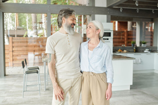 快乐住在大房子里的现代老年夫妇的画像：他们在厨房里互相拥抱室内成人成熟女人